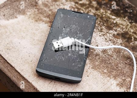 Écran tactile en verre brisé d'un smartphone noir cassé avec un câble de chargement blanc sur une surface de grunge extérieure en béton. Problèmes de communication c Banque D'Images
