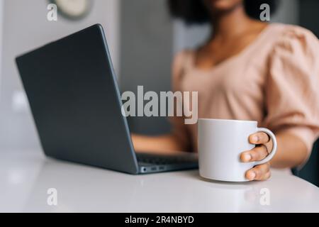 Gros plan court de femme indépendante noire non reconnaissable, dactylographiant sur un ordinateur portable et tenant une tasse de café ou de thé le matin assis Banque D'Images