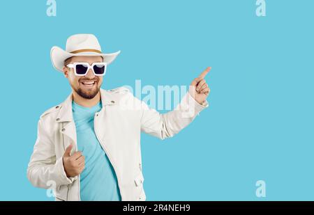Jeune homme caucasien habillé de blouson blanc et chapeau de cowboy pointe le doigt à l'espace de copie sur turquoise Banque D'Images