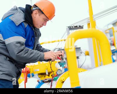 Mécanicien d'instrumentation et d'automatisation diagnostique des capteurs sur les tuyaux d'une station de distribution de gaz avec un multimètre en hiver. Inspection et diagnostic professionnels de l'état de fonctionnement des équipements à gaz. Photo d'un véritable flux de travail. Banque D'Images