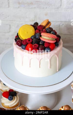 Gâteau blanc décoré d'énormes macarons, framboises, fraises et bleuets, chocolat rose fondu sur le dessus, sur le cakestan blanc Banque D'Images