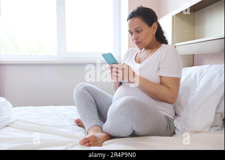 Jeune femme enceinte utilisant un smartphone, recherchant des informations sur la grossesse, vérifiant de nouvelles applications mobiles, se détend sur le lit Banque D'Images