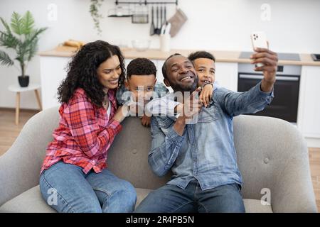 Famille positive multiculturelle de quatre personnes utilisant un smartphone moderne tout en se reposant dans le salon. Des fils adorables embrassent les parents pendant que leur père prend son selfie via une webcam mobile à l'intérieur de la chambre. Banque D'Images