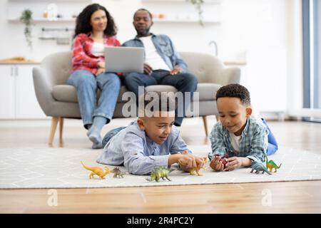 Accent sélectif sur les enfants multiculturels ludiques, couchés au sol, couverts de moquette, tandis que les conjoints décontractés sont assis sur un canapé avec ordinateur portable. Les garçons curieux étant engagés dans un jeu aventureux avec des dinosaures. Banque D'Images
