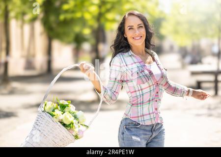 Une belle brunette heureuse danse avec joie dans les rues de la ville avec un panier rempli de fleurs fraîches. Banque D'Images