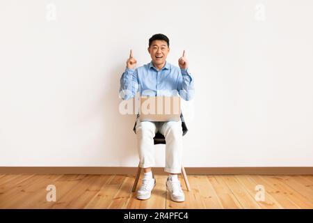 Un homme freelance d'âge moyen coréen heureux, assis sur une chaise avec un ordinateur portable de marque et pointant vers le haut sur l'espace libre au-dessus du mur lumineux Banque D'Images