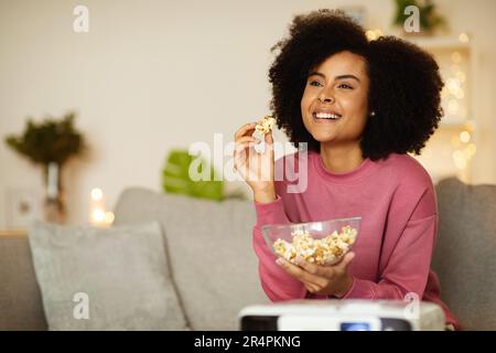 Joyeuse femme afro-américaine appréciant le tournage de films en train de manger du maïs soufflé en intérieur Banque D'Images