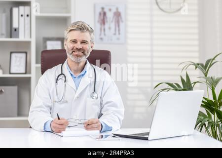 Portrait d'un médecin senior à cheveux gris, assis au bureau, à un bureau avec un carnet blanc et un stéthoscope, souriant et regardant l'appareil photo. Banque D'Images