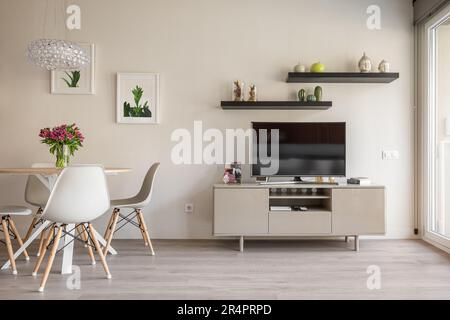 Salon confortable avec fenêtres panoramiques, table à manger et chaises en toile de fond d'un mur avec une télévision, des étagères et des accessoires décoratifs et Banque D'Images