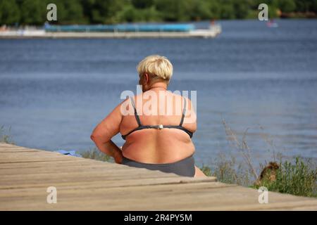 Femme en surpoids en maillot de bain assis sur fond d'eau. Vacances d'été sur la plage de la rivière, surchauffe et concept de perte de poids Banque D'Images