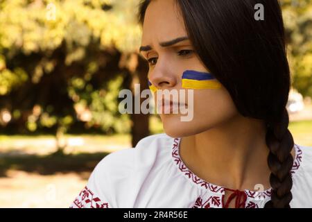 Triste jeune femme avec dessin du drapeau ukrainien sur le visage dans le parc, à proximité. Espace pour le texte Banque D'Images