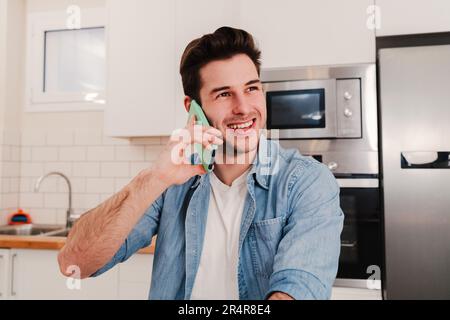 beau jeune homme appelant par téléphone portable et souriant à la cuisine maison. Homme caucasien utilisant un téléphone mobile pour avoir une conversation amicale d'appel. Je suis content de parler avec mon smartphone via une connexion sans fil. Photo de haute qualité Banque D'Images