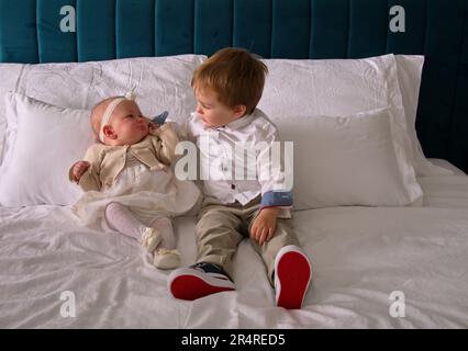 Le tout-petit et sa petite sœur sont assis sur un lit habillé pour le baptême Banque D'Images