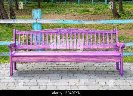 Un beau banc en bois rose se dresse sur un site pavé dans un parc, sur fond d'une clôture en bois abîmée. Banque D'Images