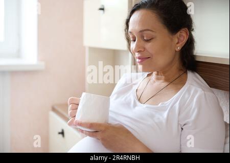 Femme enceinte avec une tasse de thé chaud ou de lait, se détendre sur le lit, profiter de sa grossesse et de son mode de vie de maternité Banque D'Images