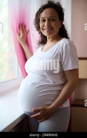 Belle femme enceinte, femme enceinte debout près de la fenêtre, touche le ventre en fin de grossesse, sourit regardant la caméra Banque D'Images