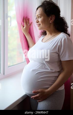 Belle femme enceinte ethnique, femme enceinte expectative regardant de rêve par la fenêtre, touchant son ventre à la fin de la grossesse Banque D'Images