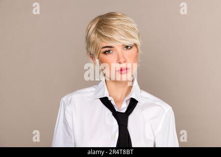 Jeune entrepreneur auto-assuré avec blond court en chemise blanche et cravate noire regardant la caméra sur fond beige Banque D'Images
