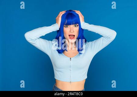 Modèle de jeune femme choqué avec des cheveux bleu vif teint dans des vêtements décontractés saisissant la tête et regardant la caméra avec la bouche ouverte en studio Banque D'Images