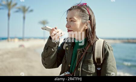 Femme hispanique mature avec cheveux gris touriste portant sac à dos parlant au téléphone au bord de la mer Banque D'Images