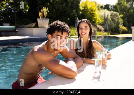 Portrait d'un jeune couple biracial joyeux tenant des boissons tout en se tenant dans la piscine Banque D'Images