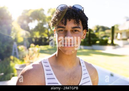 Portrait rapproché d'un jeune biracial élégant avec des lunettes de soleil se posant dans la cour contre le ciel clair Banque D'Images