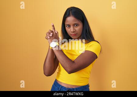 Jeune femme indienne debout sur fond jaune tenant un pistolet symbolique avec un geste de main, jouant tuant des armes de tir, visage en colère Banque D'Images