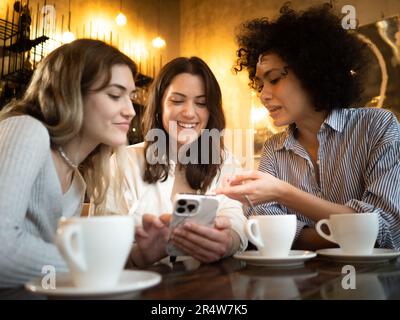 Trois jeunes femmes multiethniques se moque dans un café en train de rire et de parler tout en regardant le smartphone Banque D'Images