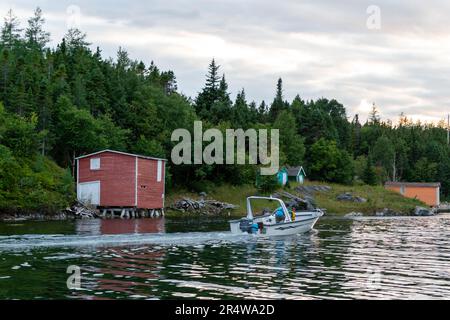 Un hangar à bateaux rouge avec une grande porte blanche sur le côté d'une rivière alors qu'un bateau à moteur remonte la rivière. La banque est couverte d'arbres verts luxuriants et d'un ciel gris. Banque D'Images