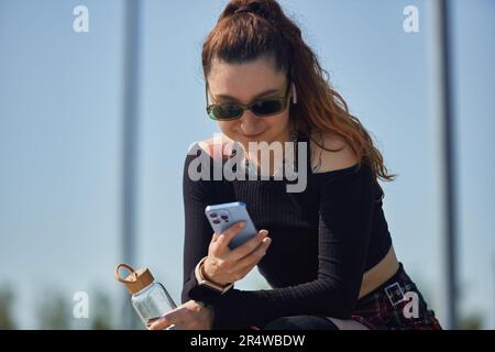 Jeune patineuse sympa, femme qui navigue sur une application mobile et tient une bouteille d'eau en verre entre les mains. Portrait du blaleur à rouleau tatoué à l'écoute de musique et de ch Banque D'Images