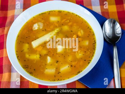 Soupe de légumes aux asperges, pommes de terre, céleri et carottes dans une assiette blanche, cuillère, serviette bleue et nappe à carreaux colorés, gros plan. Banque D'Images