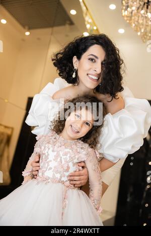 charmante mariée du moyen-orient avec cheveux brunette debout dans une robe de mariage blanche avec des manches bouffantes et des volants derrière la jolie fille et le sourire togethe Banque D'Images