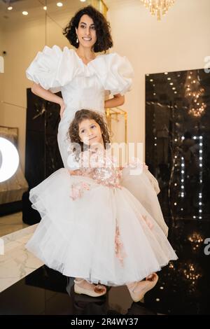 charmante mariée du moyen-orient avec des cheveux de brunette debout dans une robe de mariage blanche avec des manches de bouffée et des volants et regardant le miroir près de la tenue de fille Banque D'Images