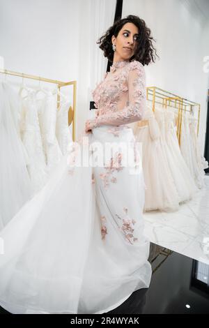 charmante et moyenne-orientale femme avec des cheveux ondulés essayant sur élégant et floral robe de mariage avec train à l'intérieur de luxueux salon de mariage, shopping, bri Banque D'Images