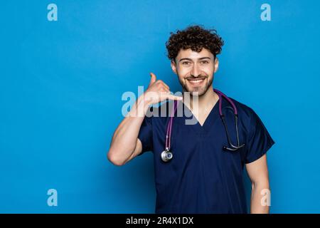 Beau médecin chauve homme avec barbe portant des lunettes et stéthoscope sur fond bleu sourire en parlant sur le geste de téléphone et de pointage t Banque D'Images