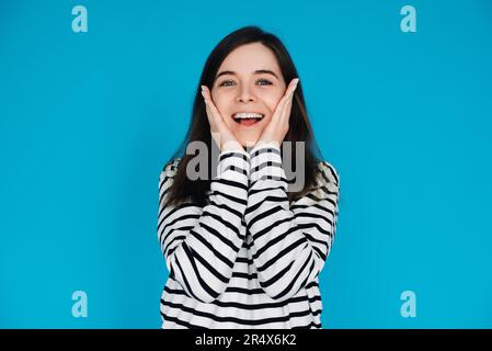 Portrait expressif d'une jeune femme surprise et joyeuse avec palmiers sur pommettes, isolé sur fond bleu. Banque D'Images