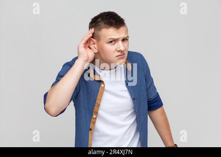 Portrait d'un adolescent portant une chemise bleue tenant la main près de l'oreille et écoutant attentivement, ayant des problèmes d'audition, la surdité dans la communication. Prise de vue en studio isolée sur fond gris. Banque D'Images