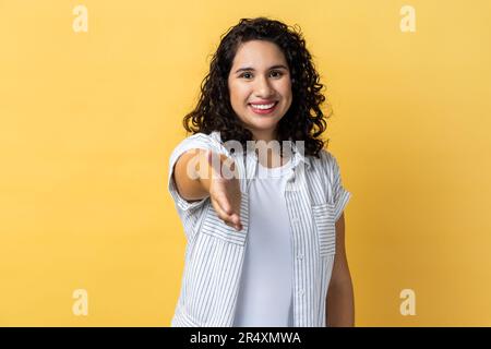 Permettez-moi de me présenter. Portrait d'une femme aux cheveux ondulés foncés femme amicale donnant la main à la poignée de main, saluant les invités avec un sourire goûteux. Studio d'intérieur isolé sur fond jaune. Banque D'Images