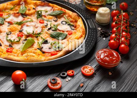Pizza avec jerky, olives, fromage et herbes sur un plat rond noir, légumes frais, sauces et épices, sur fond de bois sombre. Banque D'Images