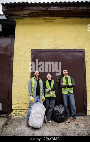 Souriant travailleurs interraciaux dans des gants et des gilets de protection regardant la caméra tout en se tenant près des sacs en plastique avec des déchets près de la porte de l'élimination des déchets Banque D'Images