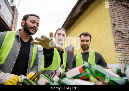 Souriant travailleurs multiethniques dans des gants et des gilets de protection regardant les déchets de verre flous dans la station d'élimination des déchets extérieure, le tri des déchets et le recyc Banque D'Images