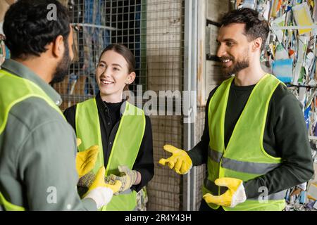 Homme gai dans un gilet et des gants de haute visibilité discutant avec des collègues multiethniques tout en se tenant et en se reposant dans un centre de tri des déchets, tri des déchets Banque D'Images