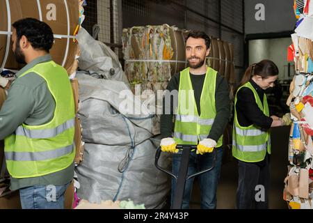 Travailleur souriant dans un gilet de protection et des gants debout près d'un transpalette manuel et à côté de collègues multiethniques travaillant avec des déchets de papier dans les ordures Banque D'Images