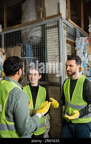 Homme souriant dans un gilet de sécurité et des gants parlant à un collègue indien tout en se reposant près du papier de rebut dans le centre de tri des déchets, le tri des déchets et Banque D'Images