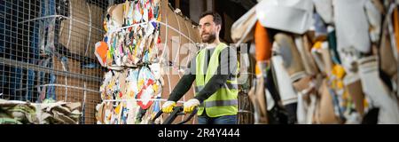 Trieur barbu dans un gilet haute visibilité et des gants se tenant près d'un transpalette tout en se tenant près du papier usagé dans un centre de tri des déchets flou, W Banque D'Images