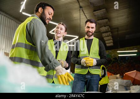 Travailleur indien dans un gilet de protection et un gant pointant vers des ordures floues et parlant à des collègues souriants et debout ensemble dans un centre de tri des ordures Banque D'Images