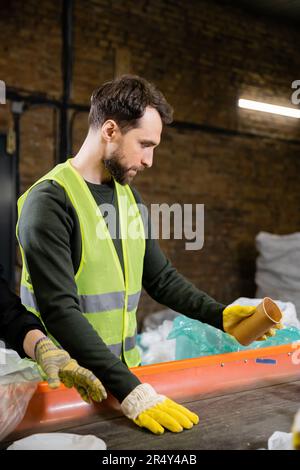 Trieur dans un gilet réfléchissant et gants de protection tenant un gobelet en papier près du convoyeur et de l'employé tout en travaillant dans une station d'élimination des déchets, en triant les déchets Banque D'Images