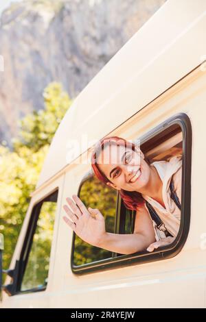 Une jeune femme agite par la fenêtre de sa camionnette de camping. Vacances en minibus et aventure en plein air en été. Concept de style de vie nomade Banque D'Images