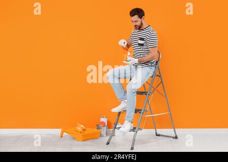 Concepteur avec équipement de peinture assis sur une échelle pliante près d'un mur orange fraîchement peint à l'intérieur, espace pour le texte Banque D'Images