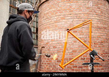 Jeune hispanique utilisant un chalumeau pour enlever la peinture d'un cadre de vélo orange qui est en cours de rénovation dans son atelier. Banque D'Images